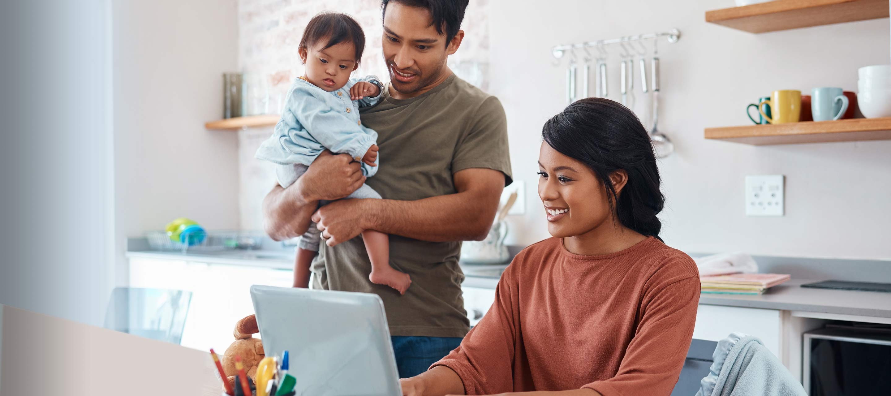 A woman is sat at the table while her husband stands next to her, holding their child. They are looking at the laptop screen.