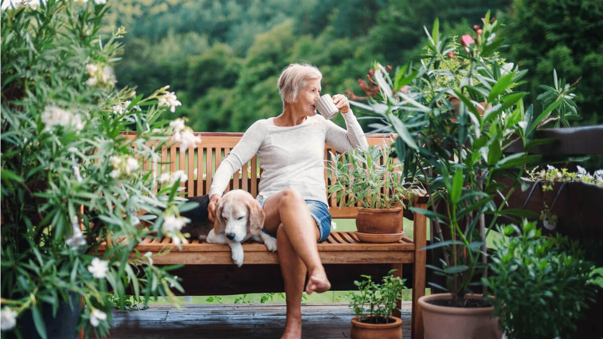 An older woman relaxes in her garden with a cup of tea and her dog.